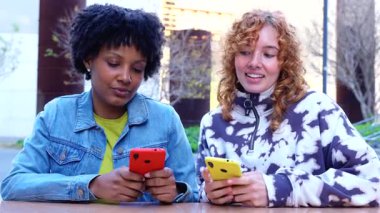 Two smiling multiracial women sitting at outdoor table using smartphones, modern lifestyle, leisure, friendship, digital technology, online communication, social connection, mobile app, young people