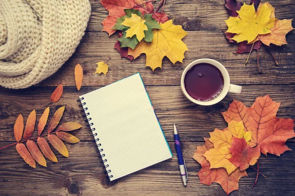 Toned picture with orange and yellow autumn leaves, warm scarf, cup of tea and Notepad with handle on wooden rustic background, top view, with copy space