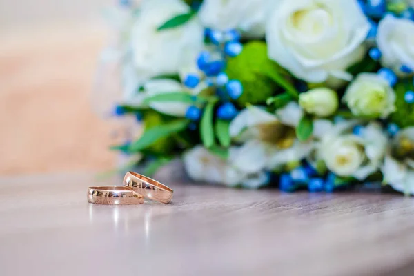 Beautiful gold wedding rings on the table next to the bride's bouquet of white and blue flowers