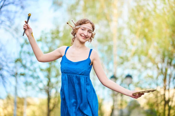 Schöne Künstlerin auf der Straße in einem blauen Kleid, lächelnd, mit Quasten und Palette in den Händen — Stockfoto