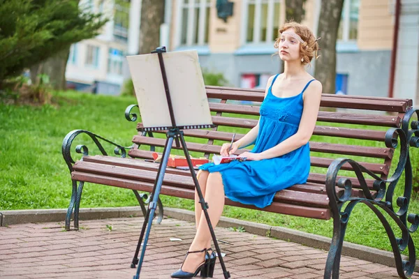 Mooi meisje-artiest op de straat in een blauwe jurk, vestigt op de ezel — Stockfoto