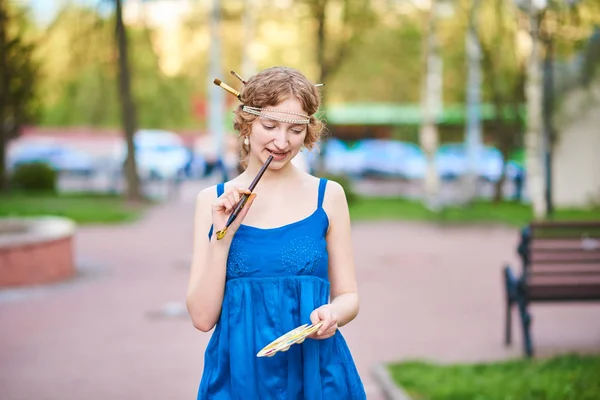 Schöne Künstlerin auf der Straße in einem blauen Kleid, lächelnd, mit Quasten und Palette in den Händen — Stockfoto