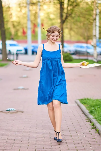 Schöne Künstlerin auf der Straße in einem blauen Kleid, lächelnd, mit Quasten und Palette in den Händen — Stockfoto
