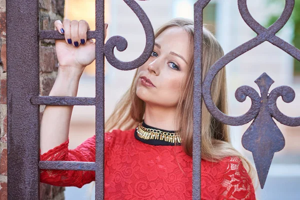 Porträtt av en vacker blond flicka i en röd klänning bakom en smidda galler — Stockfoto