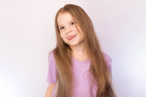Schattig klein meisje in een lavendel shirt en met haar haar, gebogen zijn hoofd naar de zijkant, toont de tong op een lichte achtergrond — Stockfoto