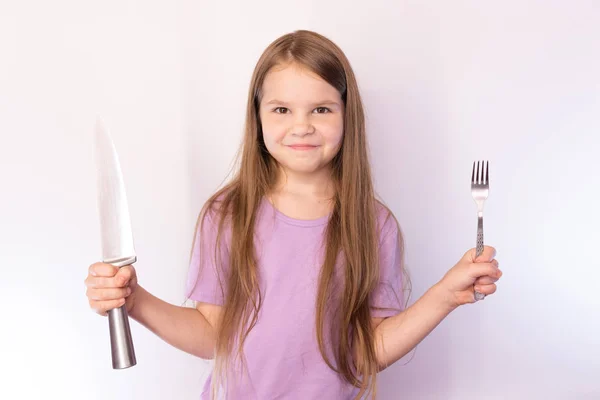 Liten söt flicka som håller en kniv och gaffel, med en nöjd uttryck på hennes ansikte, på en ljus bakgrund — Stockfoto