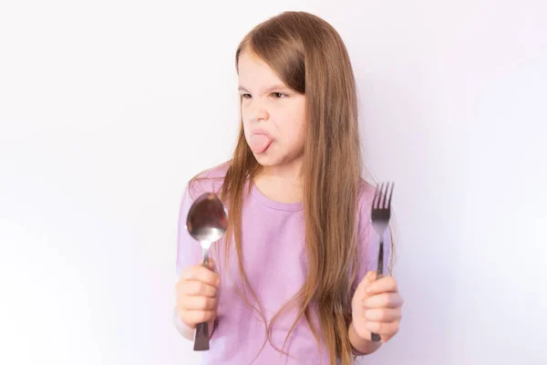 Liten söt flicka som håller en sked och gaffel i äckst sticker tungan ut och göra ett ansikte mot en ljus bakgrund — Stockfoto