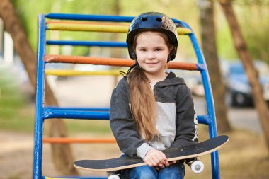 Oyun alanında kasklı küçük kız, elinde kaykay var.