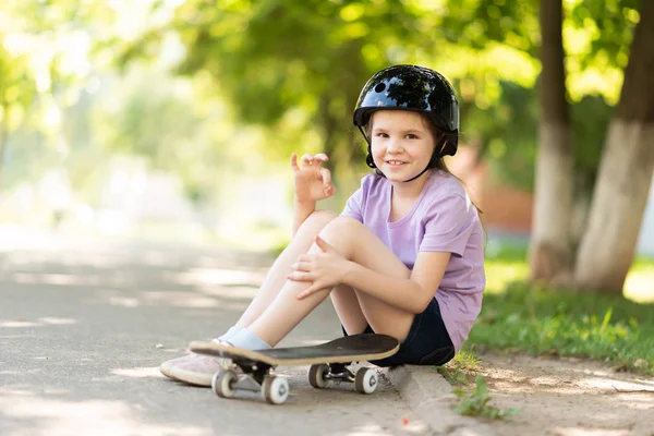 Malá holčička sedí v helmě, na skateboard a ukazuje nápis v pořádku. — Stock fotografie