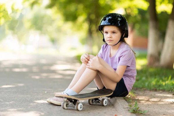 Uma menina senta-se em um skate, usando um capacete, e olha para a distância . — Fotografia de Stock