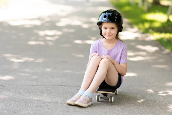 Kleines Mädchen mit Helm sitzt auf einem Skateboard. — Stockfoto
