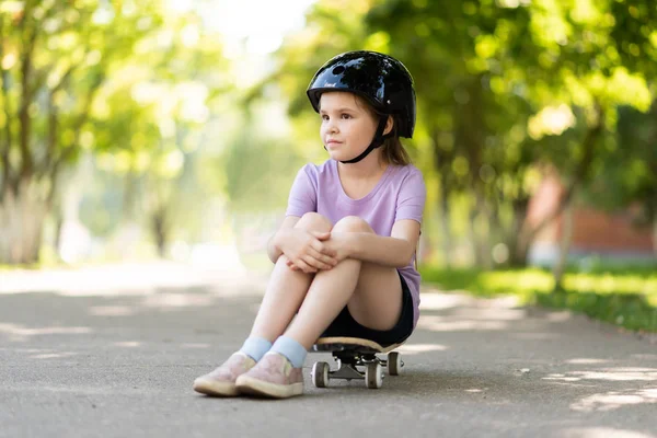 Ein kleines Mädchen mit Helm sitzt auf einem Skateboard und blickt in die Ferne. — Stockfoto