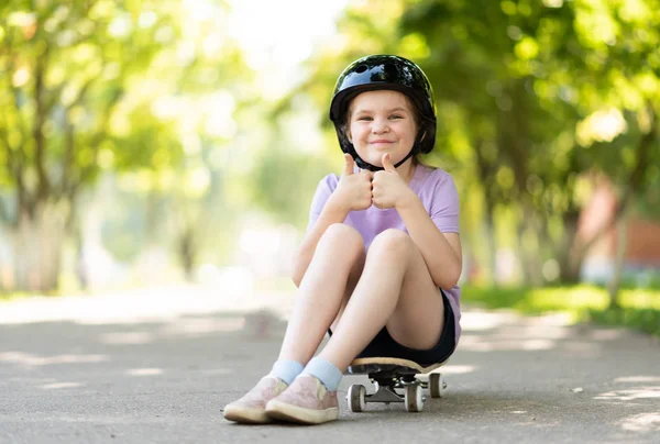 A pequena se senta em um skate, mostra um sinal de classe, com uma expressão engraçada em seu rosto . — Fotografia de Stock