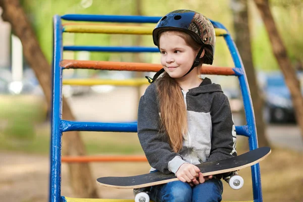 Menina em um capacete no playground, com um skate em suas mãos — Fotografia de Stock