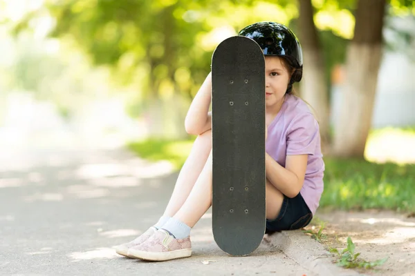 Malá holčička v helmě sedí a skrývá za skateboard. — Stock fotografie