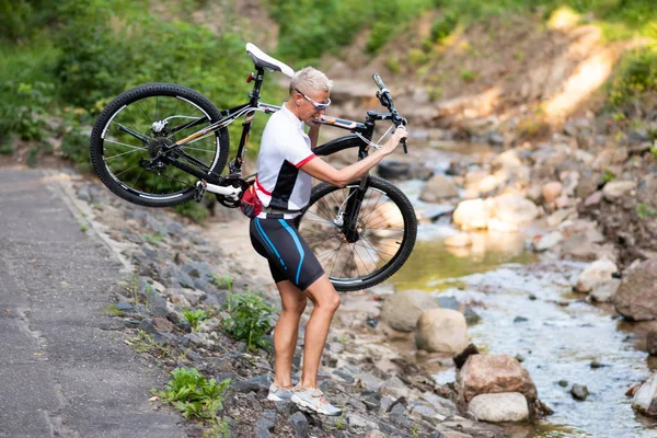 Omuzlarında bisiklet olan bir kadın derenin yanındaki taşların üzerinde duruyor..
