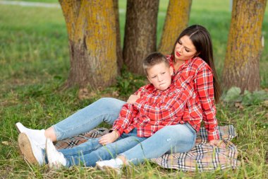 Anne ve oğlu, yatak örtüsüne yaslanıyor, oğlu kaşlarını çatıyor, dudaklarını kesiyor ve yan tarafa bakıyor..