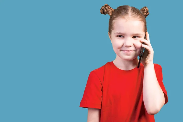 Little cute girl, in a bright T-shirt, talking on a smartphone with a slight smile on her face, isolated on a blue background.