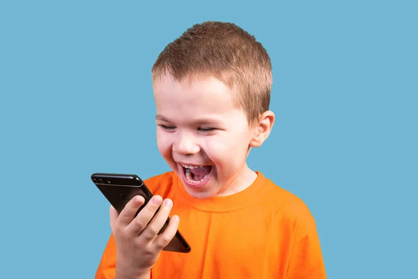 Ein kleiner Junge schaut auf das Telefon, lacht. isoliert auf blauem Hintergrund. — Stockfoto