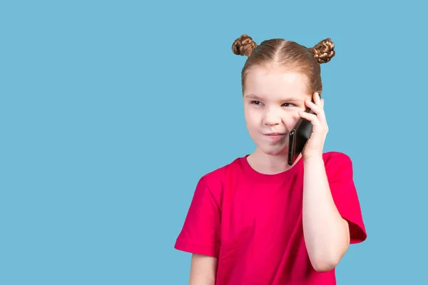 Liten söt flicka i en ljus T-shirt talar på en smartphone med en pensiv ansiktsuttryck isolerad på blå bakgrund. — Stockfoto