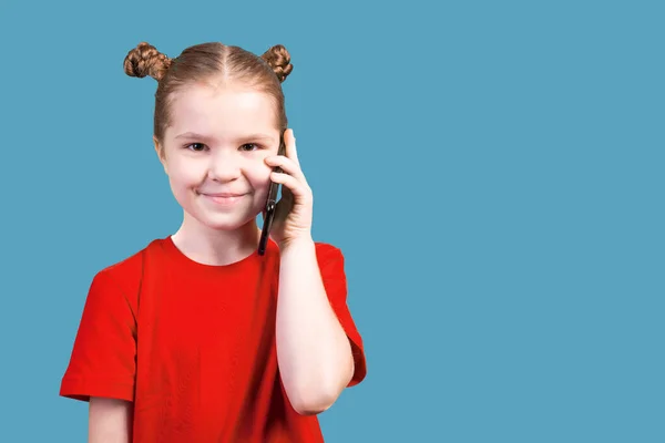 Niña linda, en una camiseta brillante, hablando en un teléfono inteligente y sonriendo, aislada sobre un fondo azul . — Foto de Stock