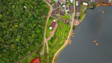 Göl kenarındaki bir köyün havadan görünüşü, kırsal mimari, yemyeşil ve sakin sulara dağılmış teknelerin karışımı sergileniyor. Doğal güzellik ve insan yerleşiminin ahenkli bir karışımı.