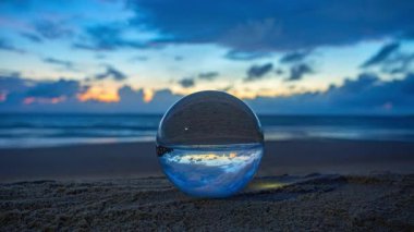 Sahile yerleştirilmiş cam bir küre, okyanusun üzerinde canlı bir günbatımının ters görüntüsünü yansıtıyor. Sıcak altın gökyüzü ile kürenin içindeki derin mavi bulutlar arasında çarpıcı bir tezat oluşturuyor.
