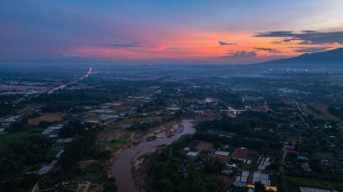 Canlı bir günbatımının nefes kesen panoramik görüntüsü, dağınık bulutlar ve uzak manzaralarla çevrili, yemyeşil bir tepenin üzerindeki gökyüzünü aydınlatıyor..