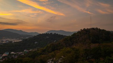Gün batımında yemyeşil dağların arasına yerleşmiş bir kasabanın manzarası. Tepenin yamacına tünemiş bir tapınak yapısı ve canlı turuncu bir gökyüzü ılık, sakin bir atmosfer yaratıyor..