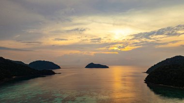Surin Adaları üzerinde çarpıcı bir günbatımının havadan görüntüsü. Sakin okyanus sularına yansıyan altın renklerle. Sahil şeridindeki mercan resifi derinlik katıyor. Şnorkel için cennet