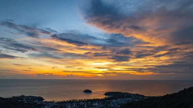 Nefes kesen bir günbatımı, kıyı kasabasının üzerinde turuncu ve mavi renkli canlı renkler çiziyor. Sahneyi çevreleyen yemyeşil tepeler var. Uzak bir tepe yapısı huzur dolu bir manzara ekler..  