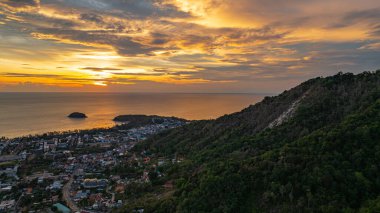 Nefes kesen bir günbatımı, kıyı kasabasının üzerinde turuncu ve mavi renkli canlı renkler çiziyor. Sahneyi çevreleyen yemyeşil tepeler var. Uzak bir tepe yapısı huzur dolu bir manzara ekler..  