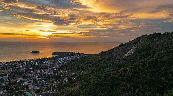 Nefes kesen bir günbatımı, kıyı kasabasının üzerinde turuncu ve mavi renkli canlı renkler çiziyor. Sahneyi çevreleyen yemyeşil tepeler var. Uzak bir tepe yapısı huzur dolu bir manzara ekler..  