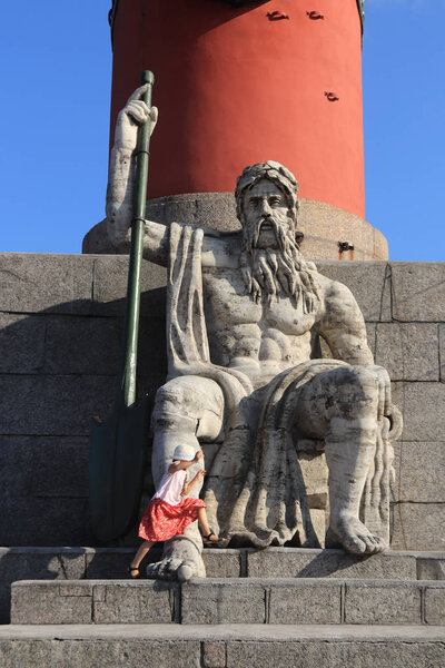 Figure of a man with a paddle sitting at the foot of the Rostral column. A little girl jumps over her right leg. St. Petersburg. Spit of Vasilyevsky Island.