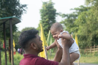 Bir baba bebeğini kucağına alır, güneşli bir açık havada göz teması kurar.