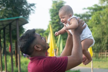 Bir adam açık havada gülümseyen bir bebeği neşeyle kaldırır.