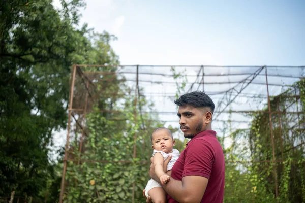 Bir adam, yeşillik ve rahat bir ortamda metal bir yapının yanında bir bebeği kucağına alır.