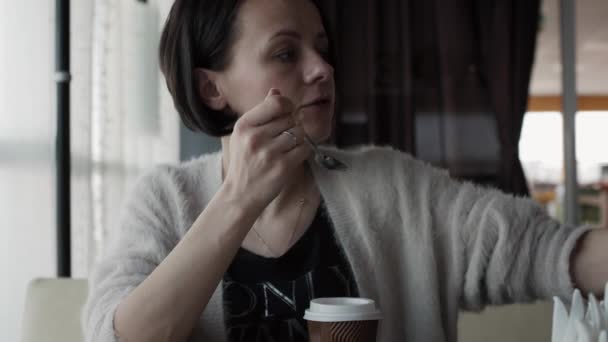 Hermosa morena comiendo pastel de queso con un smartphone en la mano — Vídeo de stock