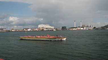 A vibrant boat tour with tourists enjoying a scenic view of an urban waterfront and industrial backdrop.