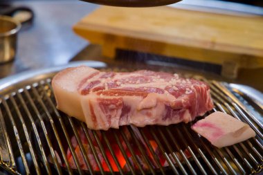 Kore barbekü ızgarasında kırmızı kömürlerin üzerinde cızırdayan Jeju 'nun yağlı tabakasına yakından bakın. Mutfak kesme tahtası ve sökücü kapağı yumuşak bir zemin oluşturuyor..