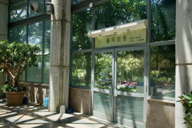 Yeomiji Botanik Bahçesi 'ndeki Sucul Bahçesi' ne giriş. Cam kapıların üzerinde Korece ve İngilizce işaretler, beton sütunlar ve üçgenlere ayrılmış sera çatısının altındaki tropikal yerleşimlerin yansımaları var..