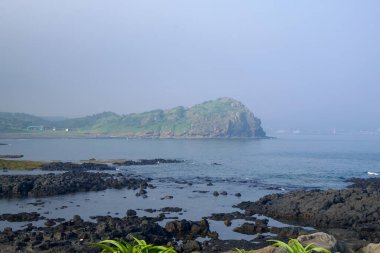 Yongmeori Sahili 'nin daha geniş bir görüntüsü, alçak yamaçlar ve kayalık gelgit düzlükleriyle desteklenen burnu gösteriyor. Bazalt kayalar ve sığ havuzlar ön planda alçak gelgitle dağılmış durumda..