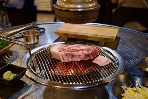 Jeju 'nun kalın bir kesimi tuz serpiştirilmiş siyah domuz eti yuvarlak bir masa ızgarasında parlayan kömürün üzerinde duruyor. Egzoz kapağı yukarıda ve muz tabakları paslanmaz masanın etrafına dizilmiş..