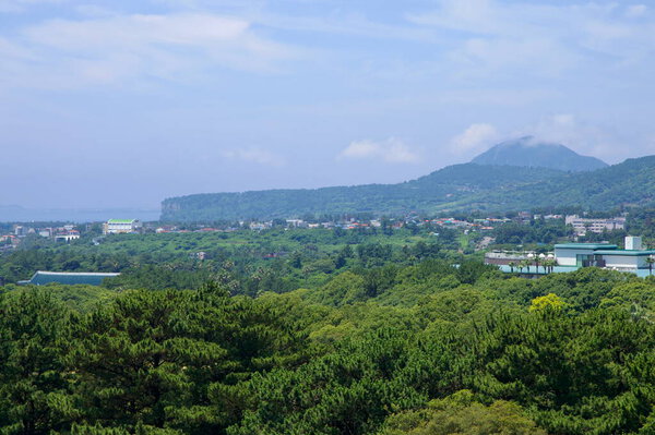 A hazy panorama from Yeomiji Botanical Garden looks toward Gunsan Oreum and the volcanic dome of Sanbangsan, with rural neighborhoods scattered across the dense forest canopy.