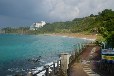 Bazalt duvarları boyunca yükselen sahil yolu Jungmun Saekdal Sahili 'ne doğru kıvrılır. Aşağıda koyu kayalar ve turkuaz su üzerinde uyarı pankartları ve korkulukları bulunur..