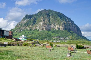 Jeju Adası 'nda arka planda yükselen Sanbangsan Dağı' nı çimenli bir sahil parkı ve ahşap pergola çerçeveleri ile süslüyor..