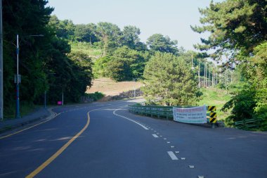 Seogwipo 'nun yeşil kenar mahallelerini gösteren, Oedolgae patikasının yakınındaki ağaçların ve inşaat afişinin yanından geçen sessiz bir yol kıvrılır..