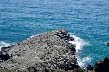 Daepo Jusangjeolli Kayalıkları 'ndaki bazalt sütunlar, derin mavi sulara karşı karanlık volkanik kayaların oluşturduğu açık deniz dalgalarıyla karşılaşıyor..