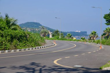 Kavisli bir yol, Beophwan yakınlarında palmiye ağaçları ve yeşillik rüzgarları ile kaplı, yol kenarından görünen taş bir haenyeo heykeli ile.