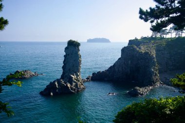 İkonik Oedolgae Kaya deniz yığını kıyı kayalıklarının ve küçük bir körfezin yanında duruyor. Bomsom Adası Seogwipo açıklarında belli belirsiz görülebiliyor..